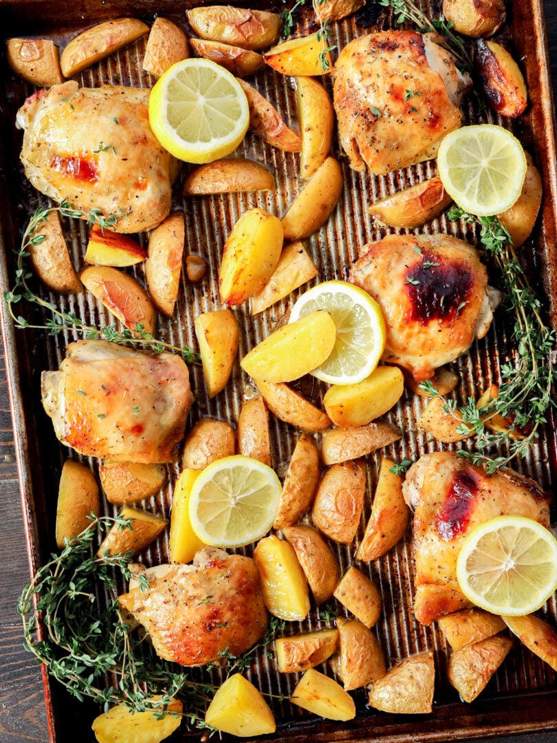 A baking sheet with Greek Lemon Chicken and Potatoes, golden wedges, lemon slices, and fresh thyme sprigs, all arranged on a striped parchment lining. The food appears well-cooked and beautifully garnished for serving.