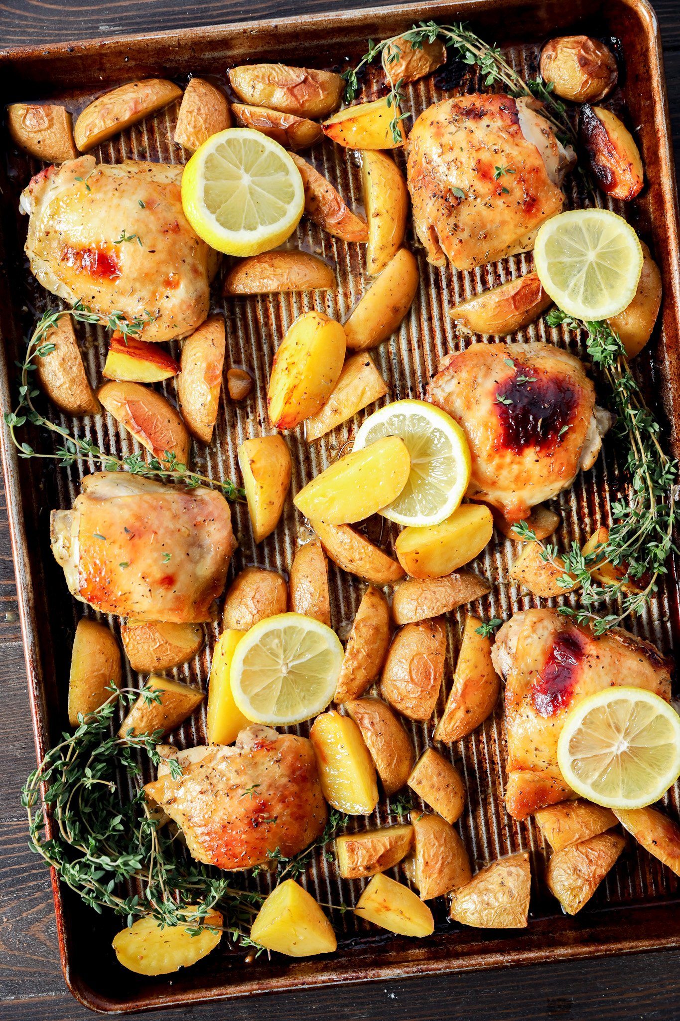 A baking sheet with Greek Lemon Chicken and Potatoes, golden wedges, lemon slices, and fresh thyme sprigs, all arranged on a striped parchment lining. The food appears well-cooked and beautifully garnished for serving.