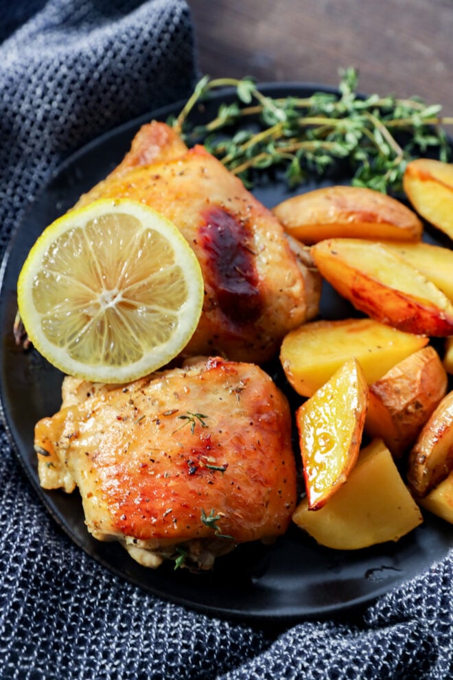 A plate of Greek Lemon Chicken and Potatoes features roasted chicken thighs, golden-brown potato wedges, a fresh lemon slice, and a sprig of herbs, all served on a dark textured fabric.