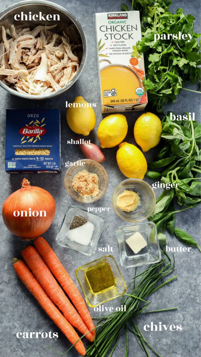 Ingredients for lemon and chicken soup are arranged on a gray surface, including shredded chicken, chicken stock, parsley, basil, lemons, shallot, garlic, ginger, orzo, onion, carrots, chives, butter, olive oil, salt and pepper. Each is labeled.