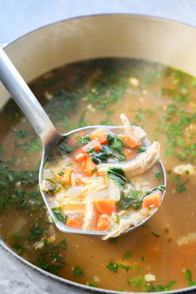 A ladle scooping homemade lemon and chicken soup with shredded chicken, diced carrots, onions, and chopped fresh herbs from a pot filled with flavorful broth.
