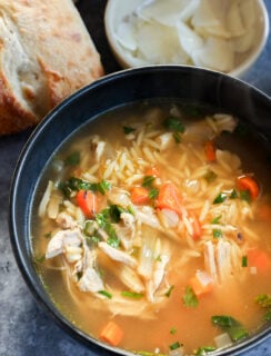A bowl of lemon and chicken soup with orzo, shredded chicken, carrots, and herbs sits next to a piece of bread and a small bowl of sliced coconut on a gray surface.