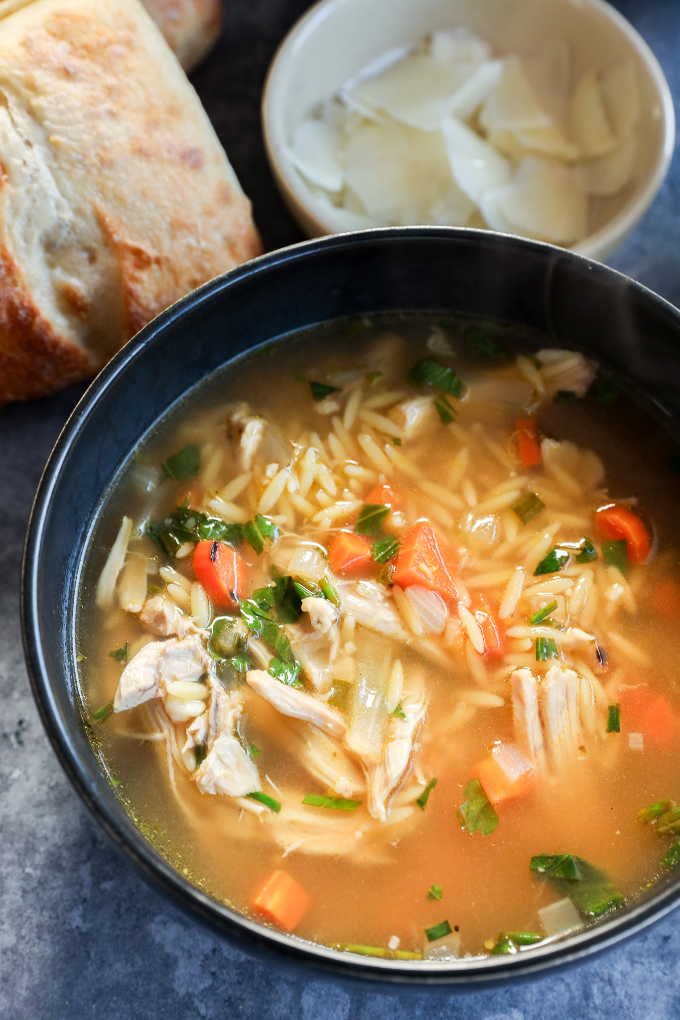 A bowl of lemon and chicken soup with orzo, shredded chicken, carrots, and herbs sits next to a piece of bread and a small bowl of sliced coconut on a gray surface.