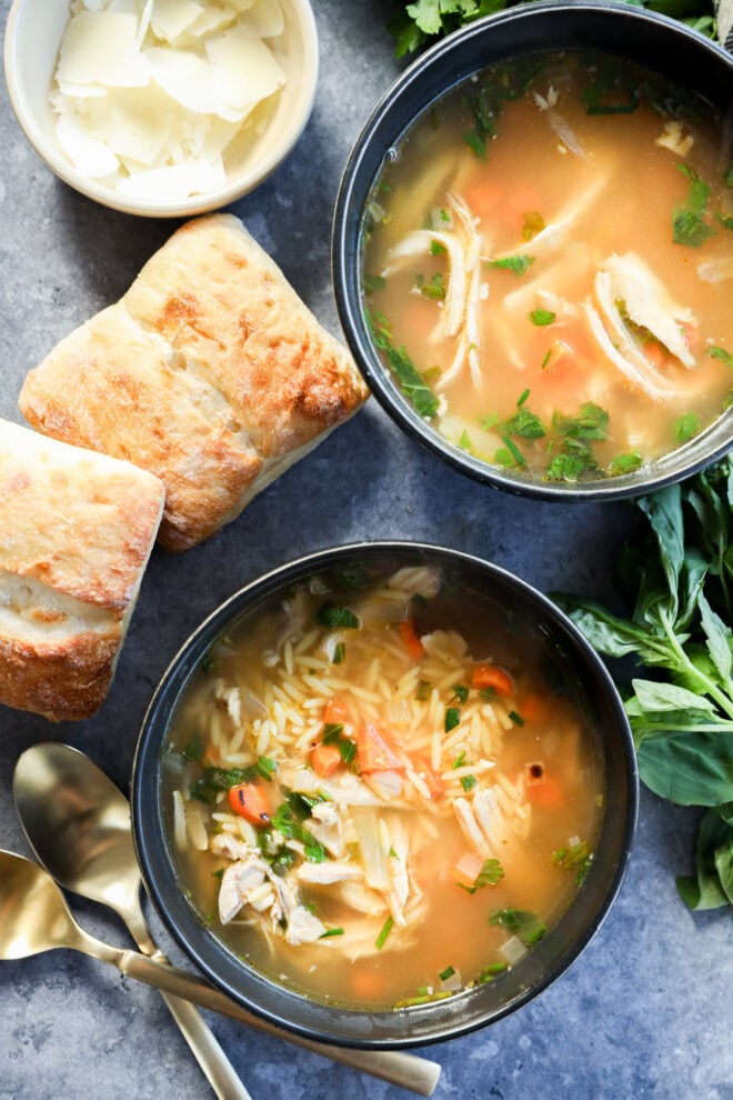 Two bowls of stew with orzo pasta and vegetables, garnished with herbs. Beside the bowls are two pieces of ciabatta, a small bowl of shaved cheese, fresh basil leaves, and two gold spoons placed nearby.