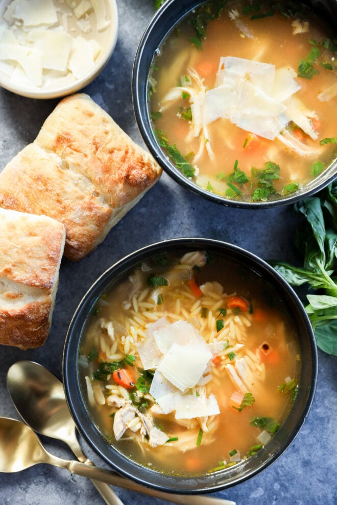 Two bowls of lemon and chicken soup with orzo, topped with shaved parmesan, are surrounded by fresh herbs, two pieces of ciabatta bread, a small bowl of parmesan, and two gold spoons on a gray surface.