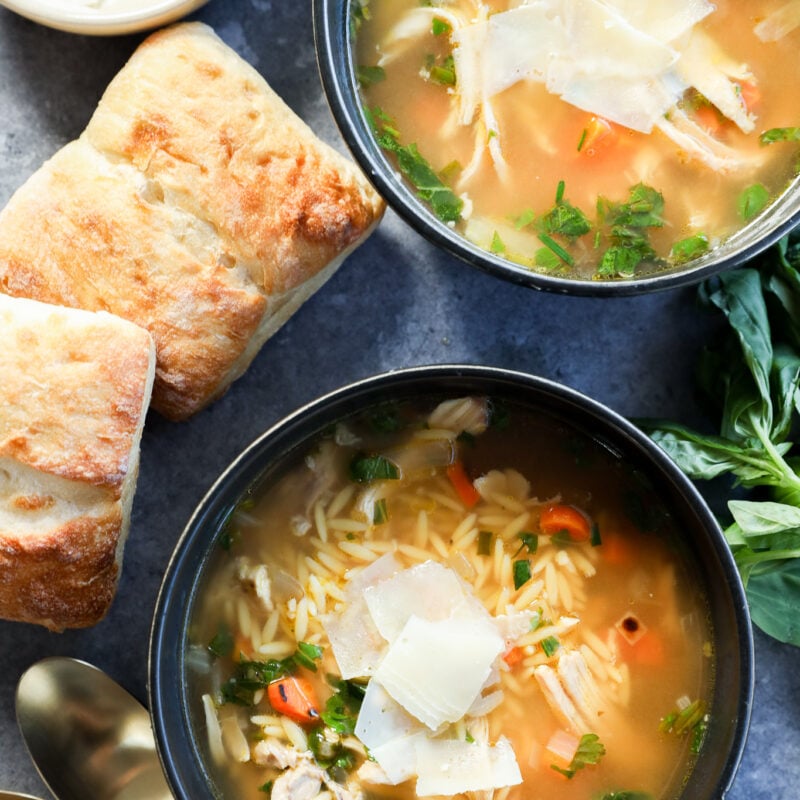 Two bowls of lemon and chicken soup with orzo, topped with shaved parmesan, are surrounded by fresh herbs, two pieces of ciabatta bread, a small bowl of parmesan, and two gold spoons on a gray surface.