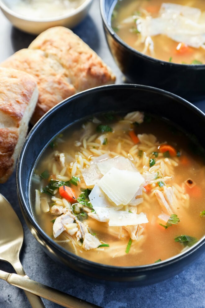 A bowl of lemon and chicken soup with orzo, garnished with shaved Parmesan cheese, sits beside a sliced baguette and another bowl in the background, with a gold spoon on the side.