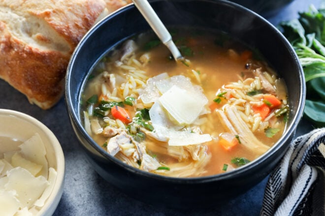 A bowl of lemon and chicken soup with orzo pasta, topped with shaved Parmesan cheese. A spoon rests inside the bowl, with crusty bread and greens visible in the background.