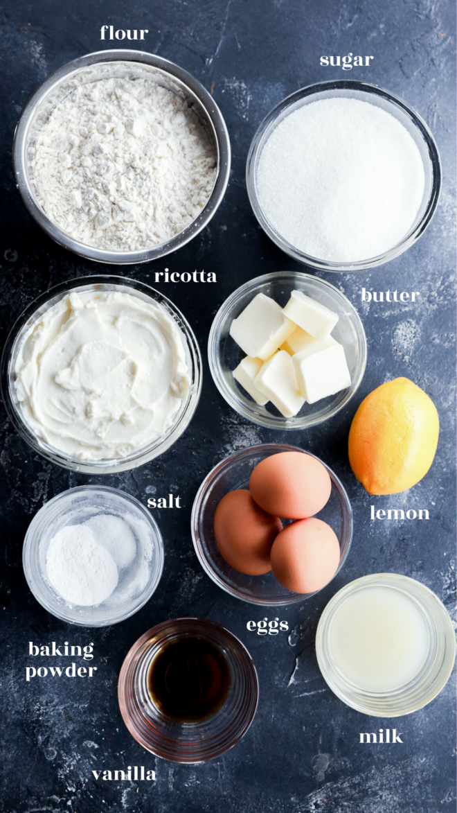 Top-down view of baking ingredients for lemon ricotta cake in bowls on a dark surface, including flour, sugar, ricotta, butter cubes, lemon, baking powder, salt, eggs, vanilla, and milk&mdash;all labeled with white text.