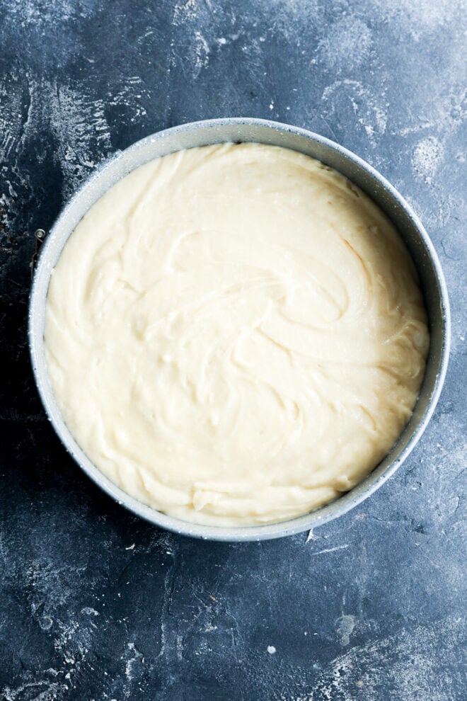 A round metal cake pan filled with smooth, pale batter, set on a dark, textured surface.
