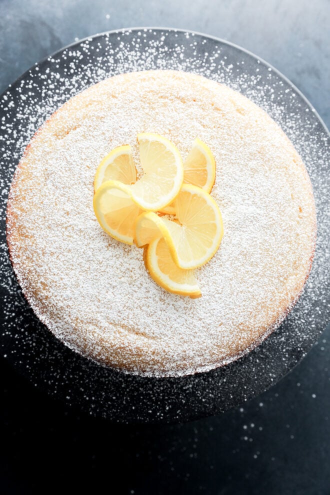 A round lemon ricotta cake dusted with powdered sugar, topped with several lemon slices, displayed on a dark plate against a dark background.