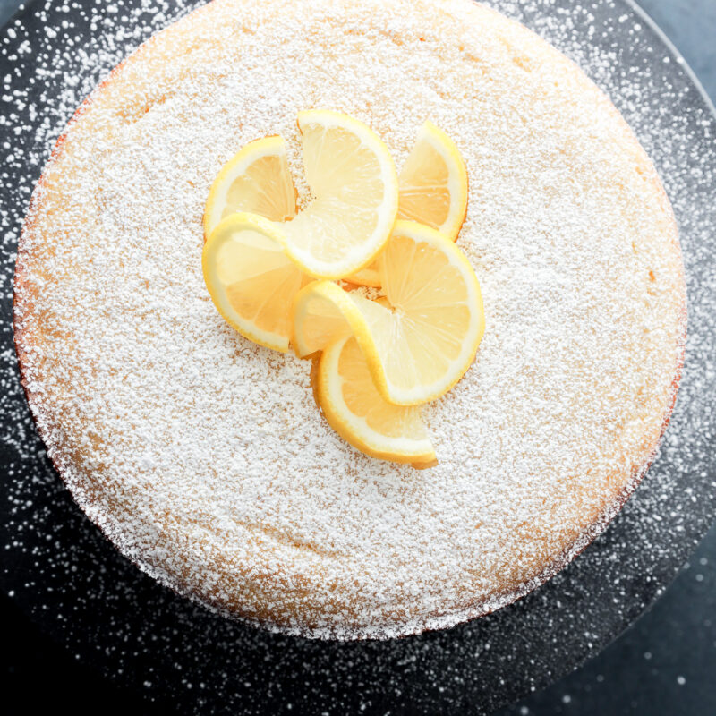 A round lemon ricotta cake dusted with powdered sugar, topped with several lemon slices, displayed on a dark plate against a dark background.