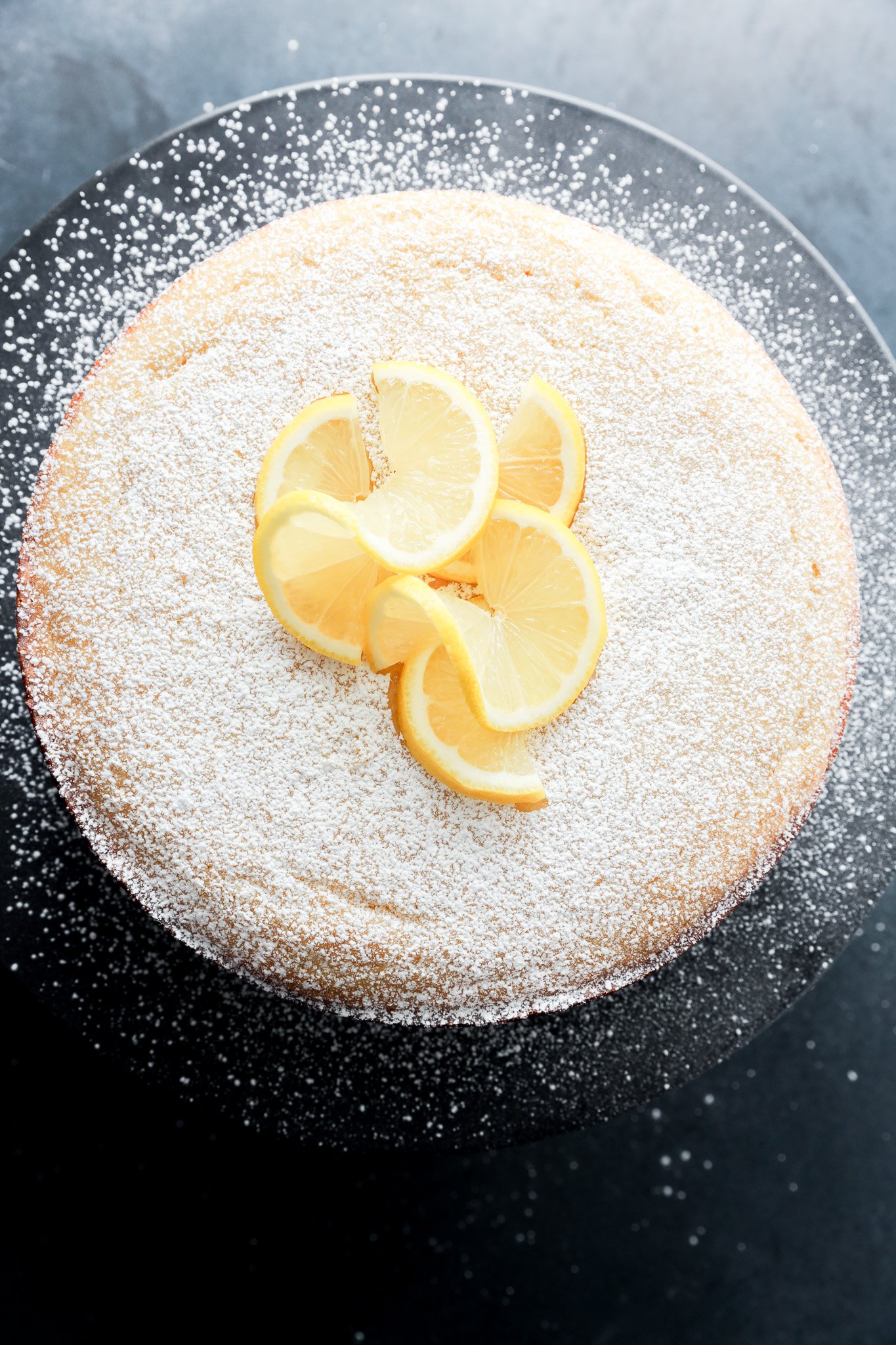 A round lemon ricotta cake dusted with powdered sugar, topped with several lemon slices, displayed on a dark plate against a dark background.
