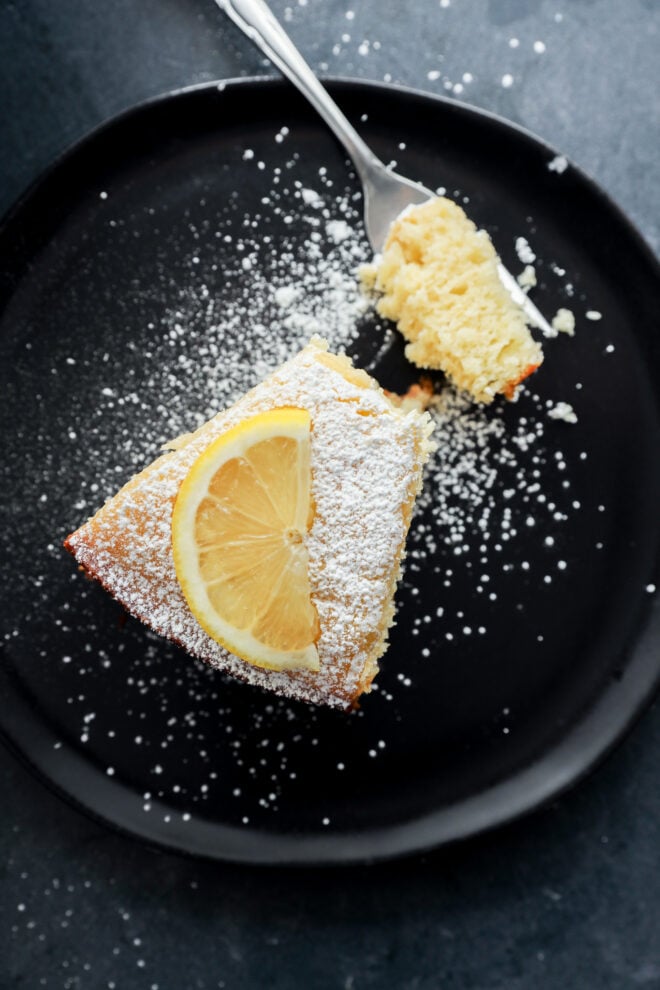 A slice of lemon ricotta cake topped with powdered sugar and a lemon slice sits on a black plate, with a fork holding a bite-sized piece nearby.