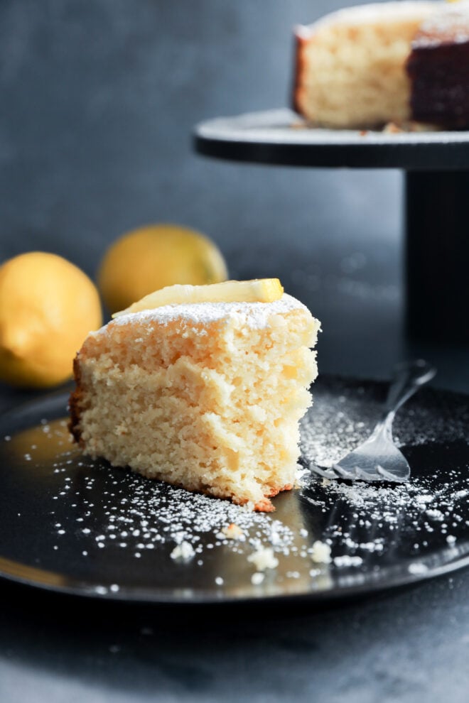 A slice of lemon ricotta cake topped with powdered sugar and a lemon wedge sits on a black plate with a fork, surrounded by whole lemons and more cake in the background.