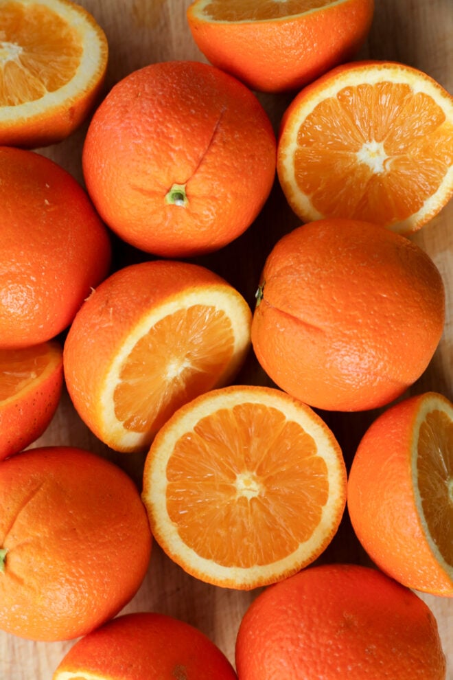 A close-up of several fresh oranges, some whole and some cut in half, revealing their juicy, bright orange flesh arranged on a wooden surface.