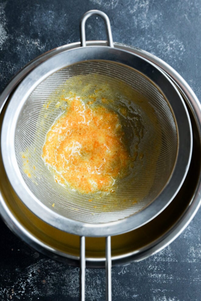 A fine mesh strainer containing orange and yellow fruit pulp is placed over a metal bowl, with fresh orange curd juice collected below and pulp left behind in the strainer. The background is a dark, textured surface.