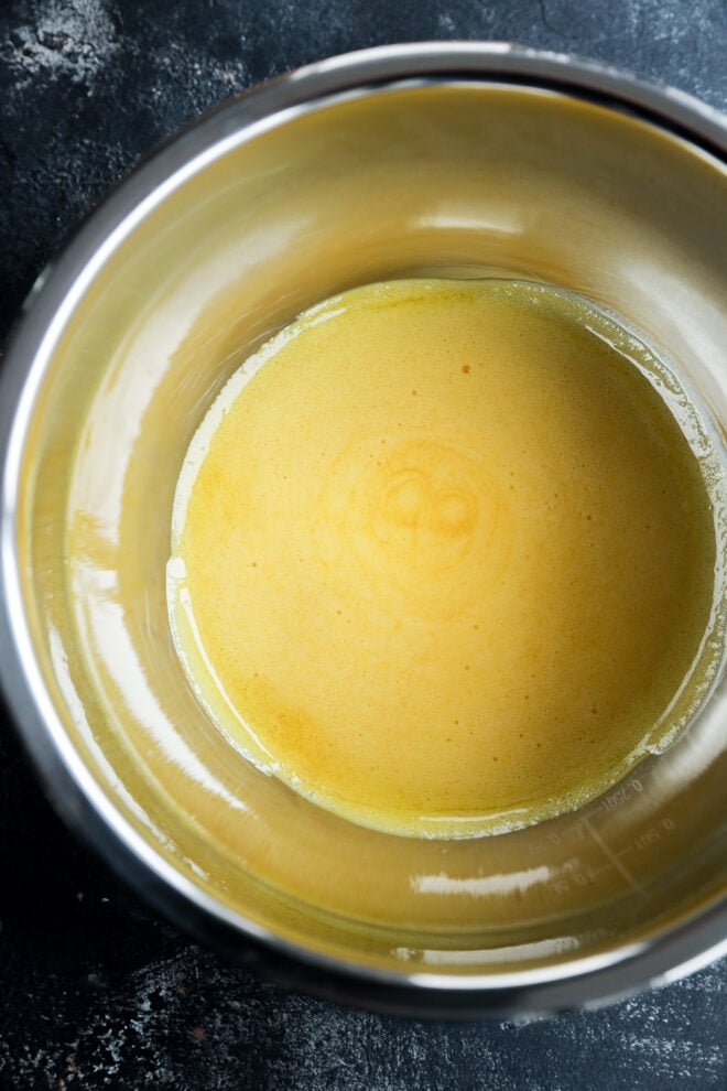 A metal mixing bowl containing a smooth, yellow orange curd mixture, viewed from above against a dark textured background.
