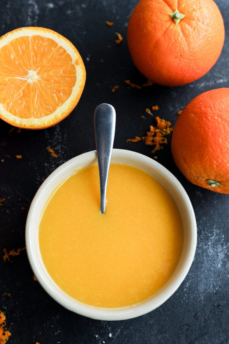 A bowl of orange curd with a spoon in it, surrounded by two whole oranges and one half orange on a dark surface, with some orange zest scattered around.