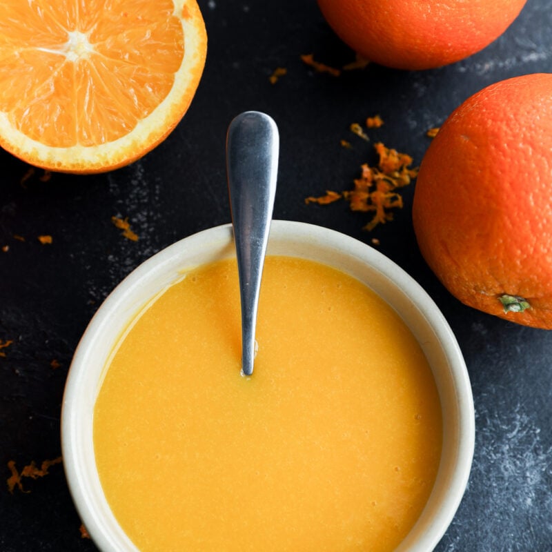 A bowl of orange curd with a spoon in it, surrounded by two whole oranges and one half orange on a dark surface, with some orange zest scattered around.