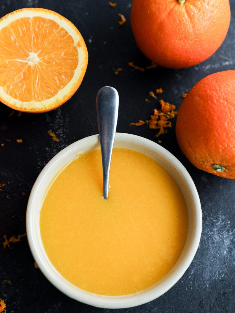 A bowl of orange curd with a spoon in it, surrounded by two whole oranges and one half orange on a dark surface, with some orange zest scattered around.