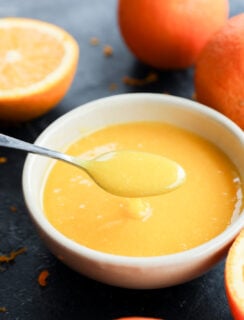 A spoon holds up a creamy orange curd above a white bowl, surrounded by fresh oranges on a dark surface.