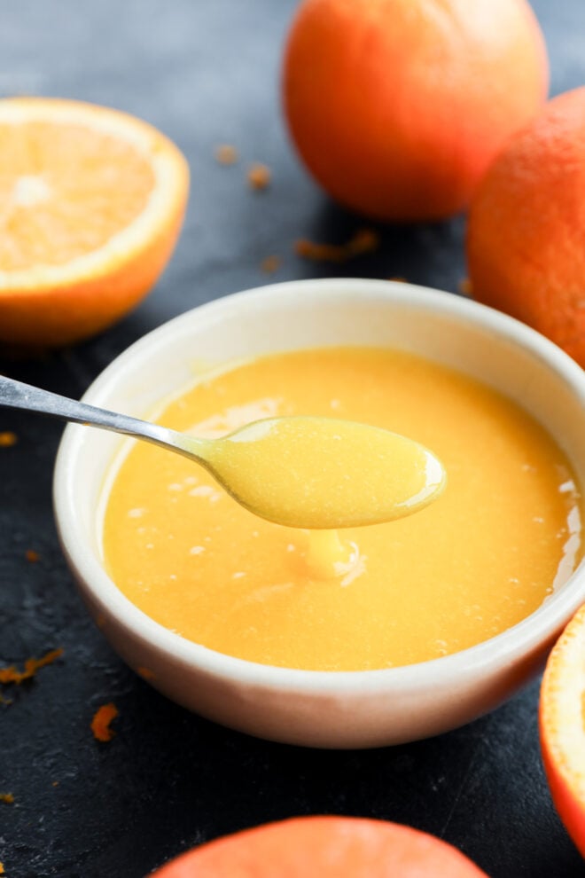 A spoon holds up a creamy orange curd above a white bowl, surrounded by fresh oranges on a dark surface.
