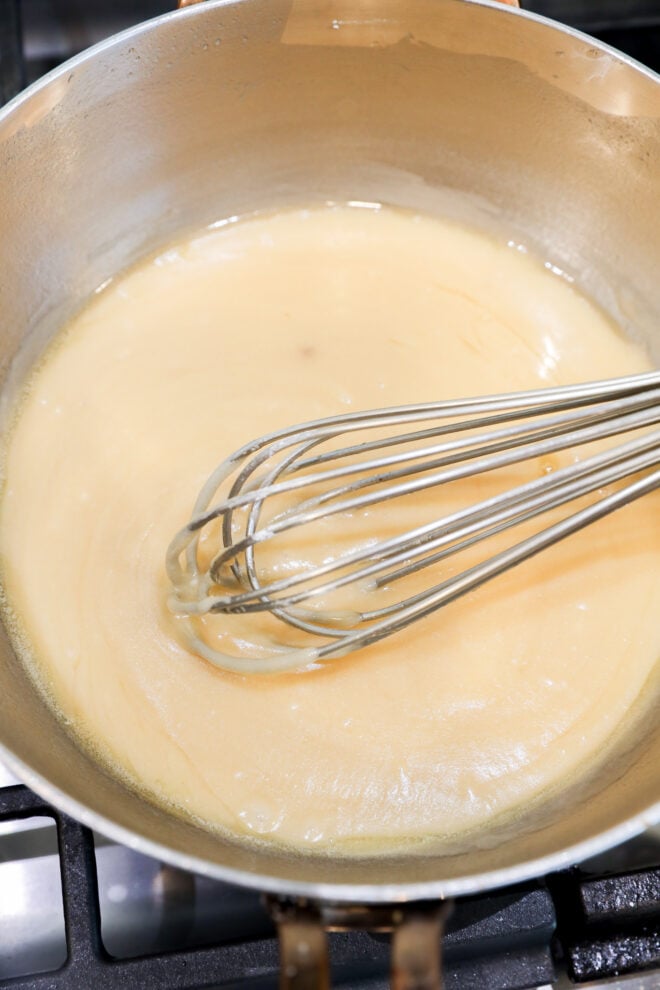 A metal whisk stirs a light tan roux in a saucepan on the stovetop, creating a smooth, slightly glossy mixture