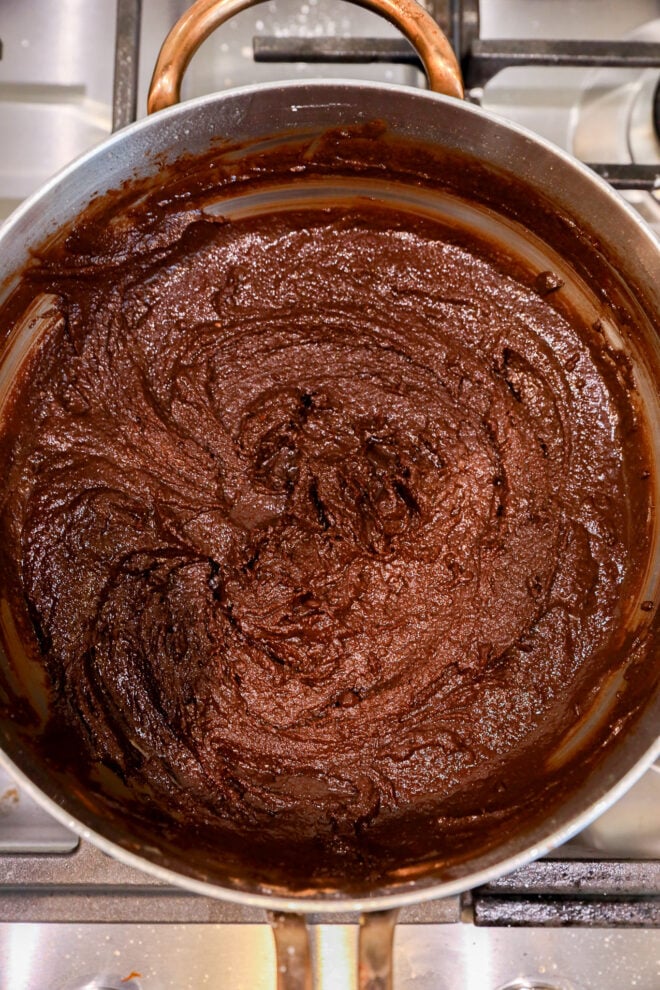 A pot on a stovetop filled with thick, dark brown chocolate mixture being stirred. The glossy texture and swirling patterns