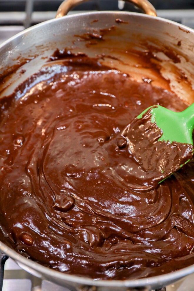 Thick, glossy chocolate brownie bites batter is being stirred in a saucepan with a green spatula on a stovetop.