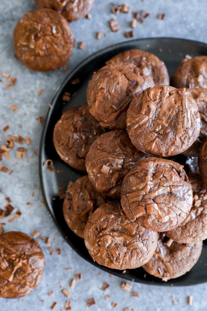 A black plate stacked with fudgy chocolate cookies and Brownie Bites, topped with chocolate shavings. More cookies and shavings are scattered on the gray surface around the plate.