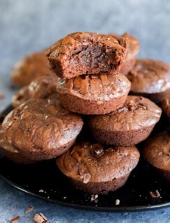 A stack of rich, fudgy chocolate brownies is displayed on a black plate. The top brownie bite has a chunk missing, revealing its moist, gooey center. Chocolate chunks are scattered on the plate and surface nearby.
