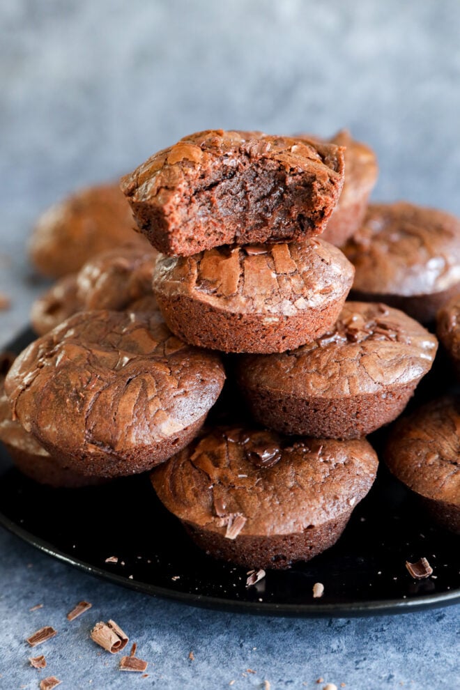 A stack of rich, fudgy chocolate brownies is displayed on a black plate. The top brownie bite has a chunk missing, revealing its moist, gooey center. Chocolate chunks are scattered on the plate and surface nearby.