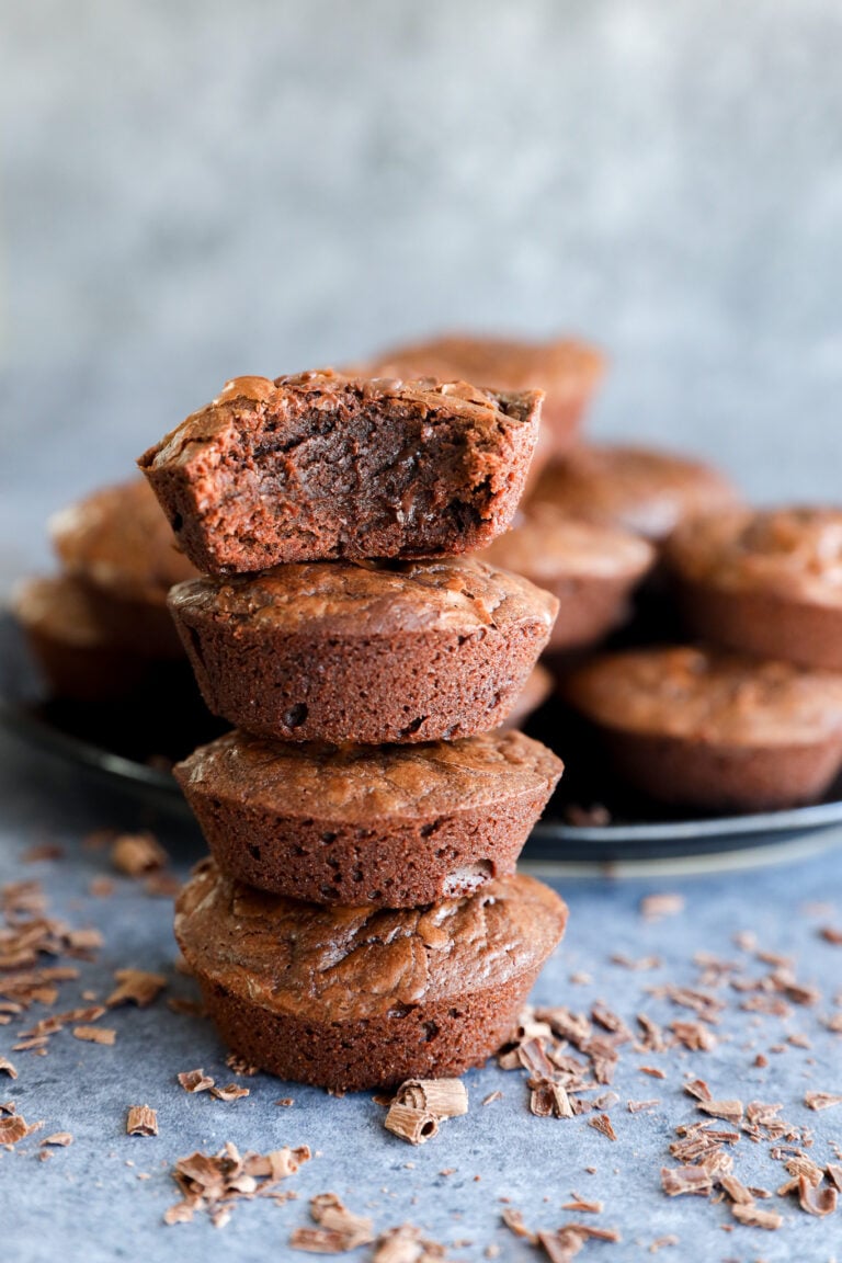 A stack of three chocolate brownies, with the top brownie missing a bite, showcases irresistible Brownie Bites. More brownies and chocolate shavings are scattered in the background on a gray surface.