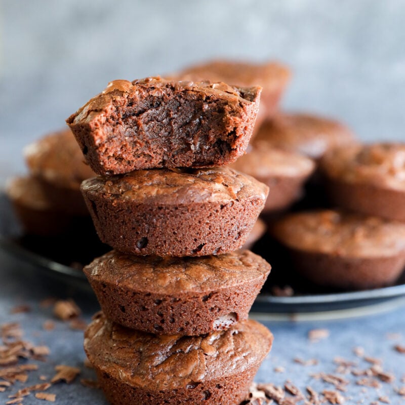 A stack of three chocolate brownies, with the top brownie missing a bite, showcases irresistible Brownie Bites. More brownies and chocolate shavings are scattered in the background on a gray surface.