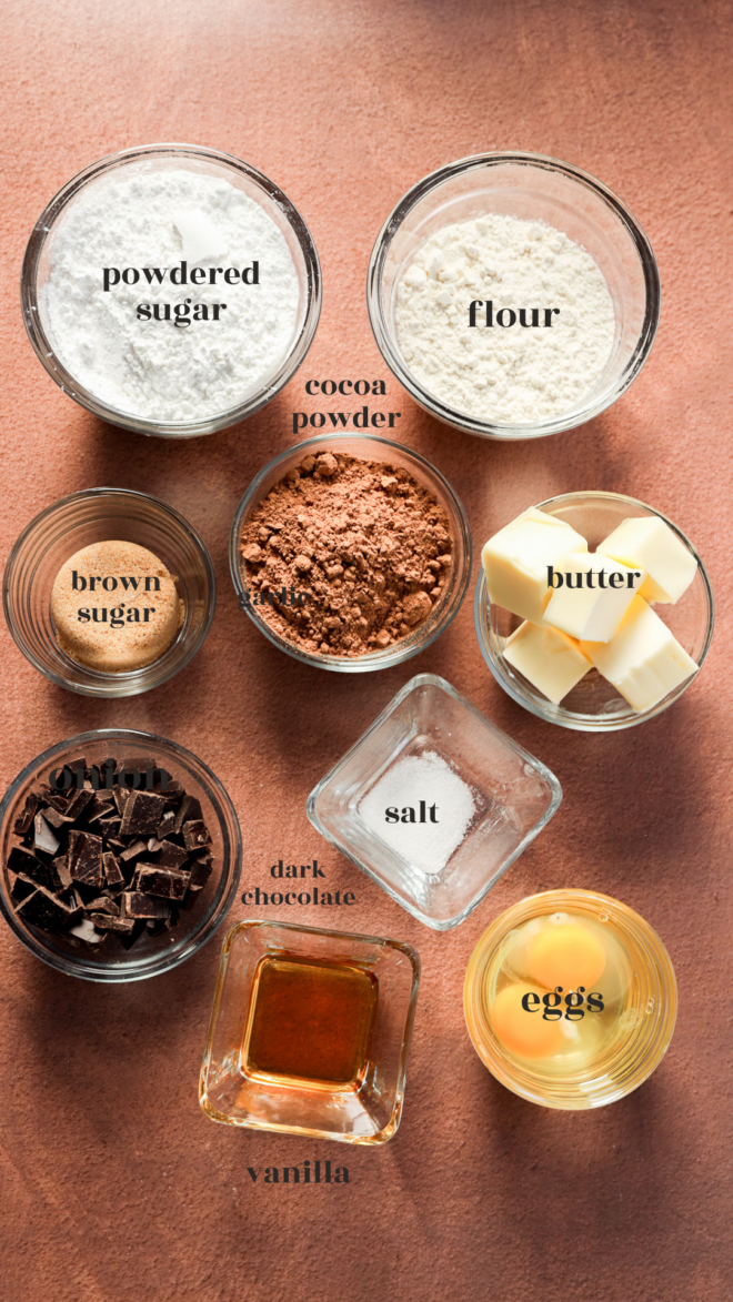 Overhead view of labeled baking ingredients in glass bowls for Brownie Bites: powdered sugar, flour, brown sugar, cocoa powder, butter, salt, dark chocolate chunks, vanilla extract, and eggs on a brown surface.