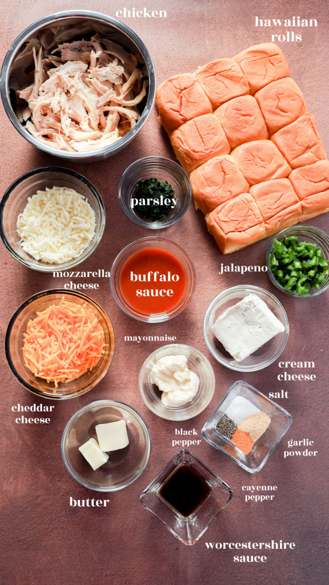 Overhead view of ingredients for buffalo chicken sliders, including shredded chicken, Hawaiian rolls, parsley, jalapeño, mozzarella, cheddar, mayonnaise, buffalo sauce, cream cheese, butter, spices, and Worcestershire sauce.