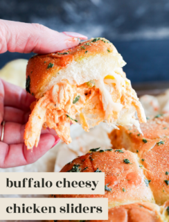 A hand holding one of the buffalo chicken sliders, with shredded chicken and melted cheese inside a golden, herb-topped bun. Other sliders rest on parchment paper below. Text on image reads: buffalo cheesy chicken sliders.