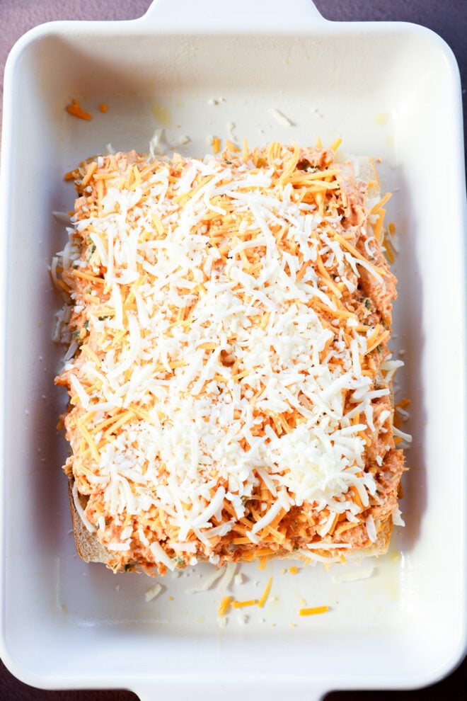 A white baking dish containing shredded chicken mixed with sauce, layered on flat tortillas like buffalo chicken sliders, and topped with a generous amount of shredded cheese, ready to be baked.