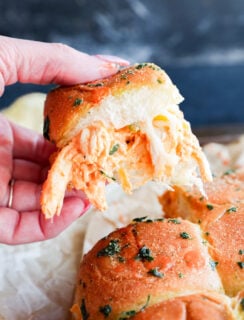 A hand holds a pulled-apart buffalo chicken slider with shredded chicken and melted cheese inside a golden, herb-topped bun. Other cheesy sliders are visible below on parchment paper.