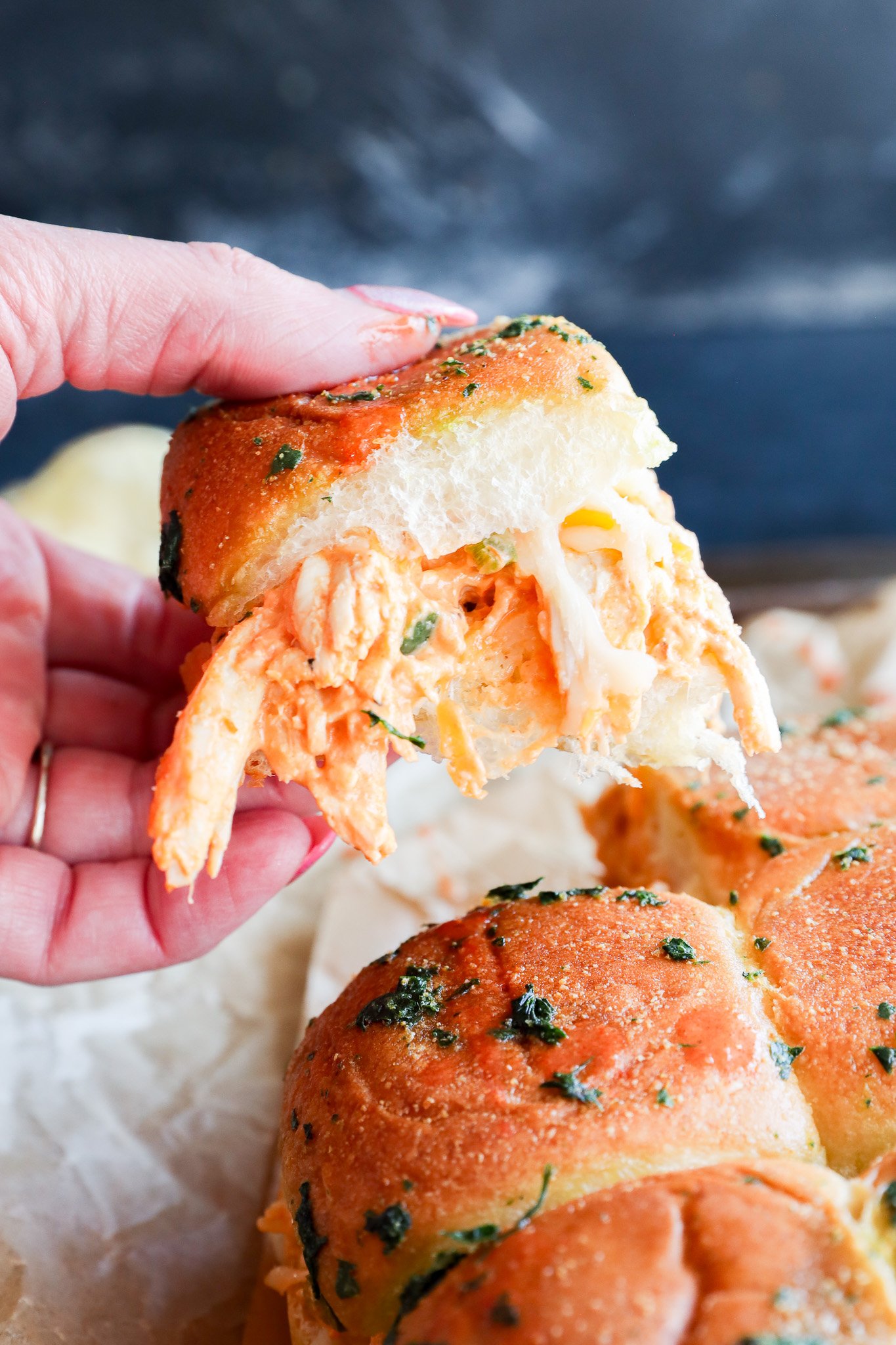 A hand holds a pulled-apart buffalo chicken slider with shredded chicken and melted cheese inside a golden, herb-topped bun. Other cheesy sliders are visible below on parchment paper.