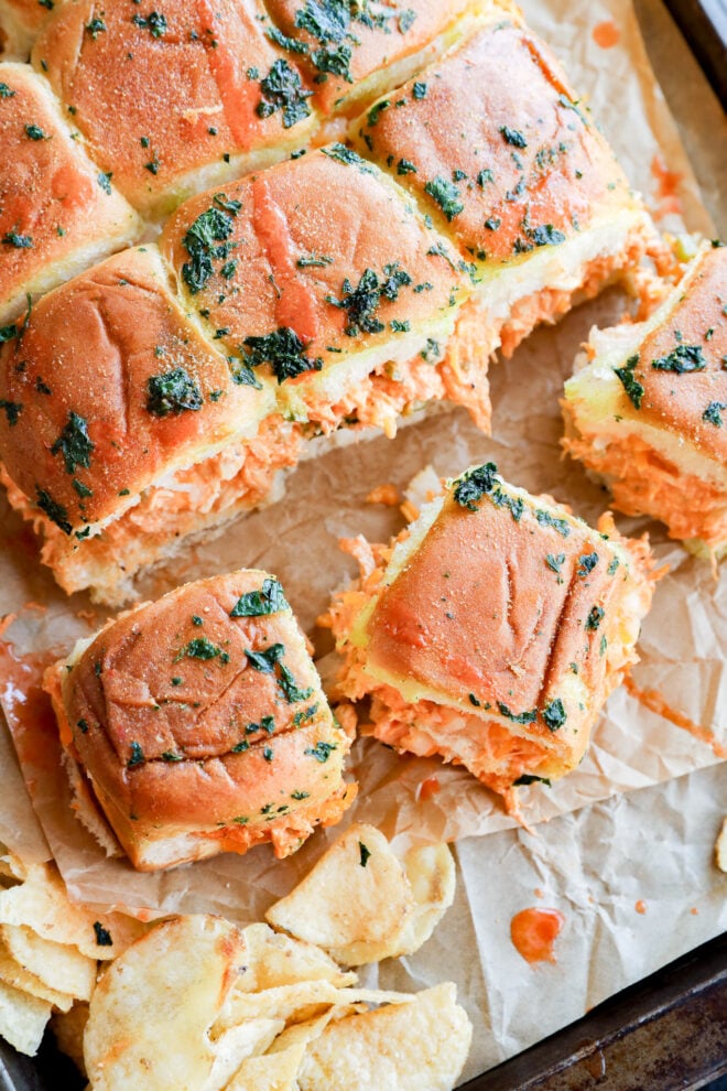 Buffalo chicken sliders with golden, herb-topped buns filled with shredded buffalo chicken sit on parchment paper, surrounded by scattered potato chips. A few sliders are pulled apart from the main group, revealing their spicy filling inside.