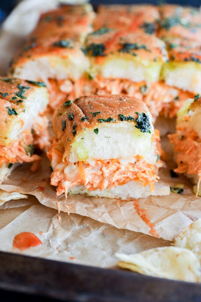 A close-up of fluffy pull-apart buffalo chicken sliders filled with shredded chicken, creamy orange sauce, and topped with herbs, set on parchment paper with potato chips beside them.
