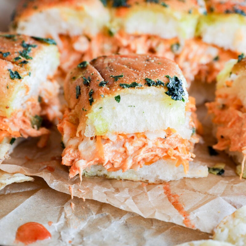 A close-up of fluffy pull-apart buffalo chicken sliders filled with shredded chicken, creamy orange sauce, and topped with herbs, set on parchment paper with potato chips beside them.