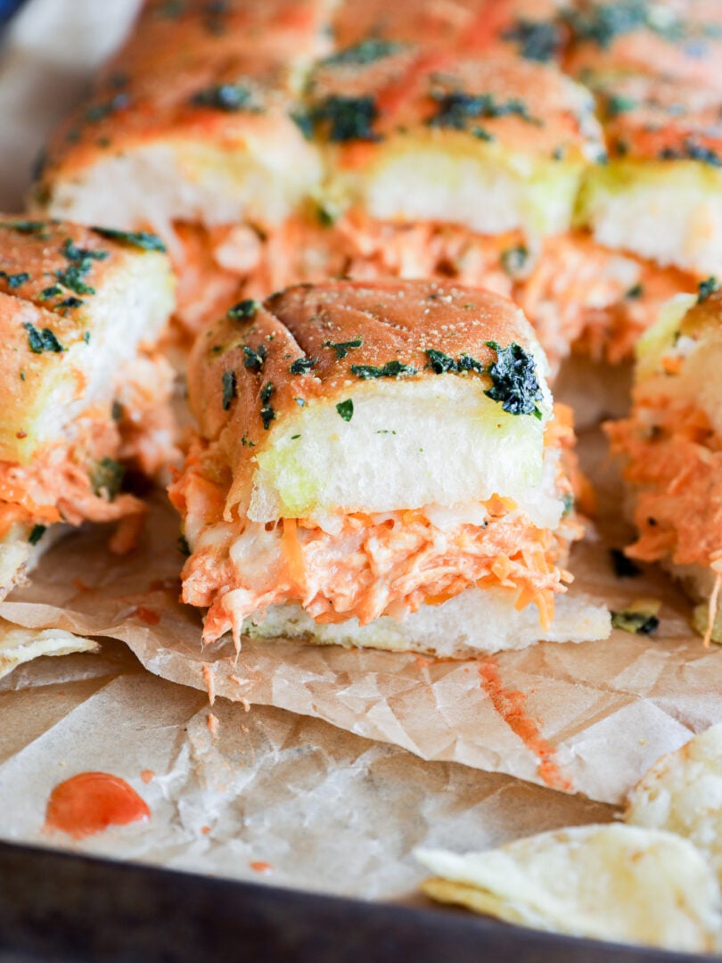 A close-up of fluffy pull-apart buffalo chicken sliders filled with shredded chicken, creamy orange sauce, and topped with herbs, set on parchment paper with potato chips beside them.