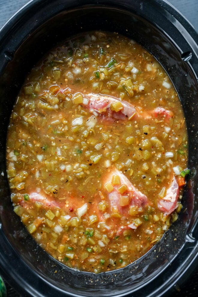 Raw pork chops are coated in a chunky pork green chile salsa with diced vegetables, all nestled in a black slow cooker and ready to be cooked.