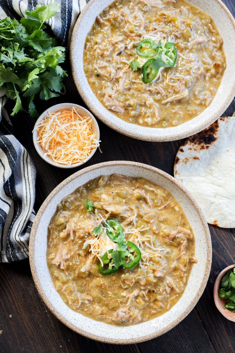 Two bowls of pork green chile stew topped with shredded cheese and sliced jalapeños, surrounded by cilantro, extra cheese, a striped towel, and a tortilla on a dark wooden table.