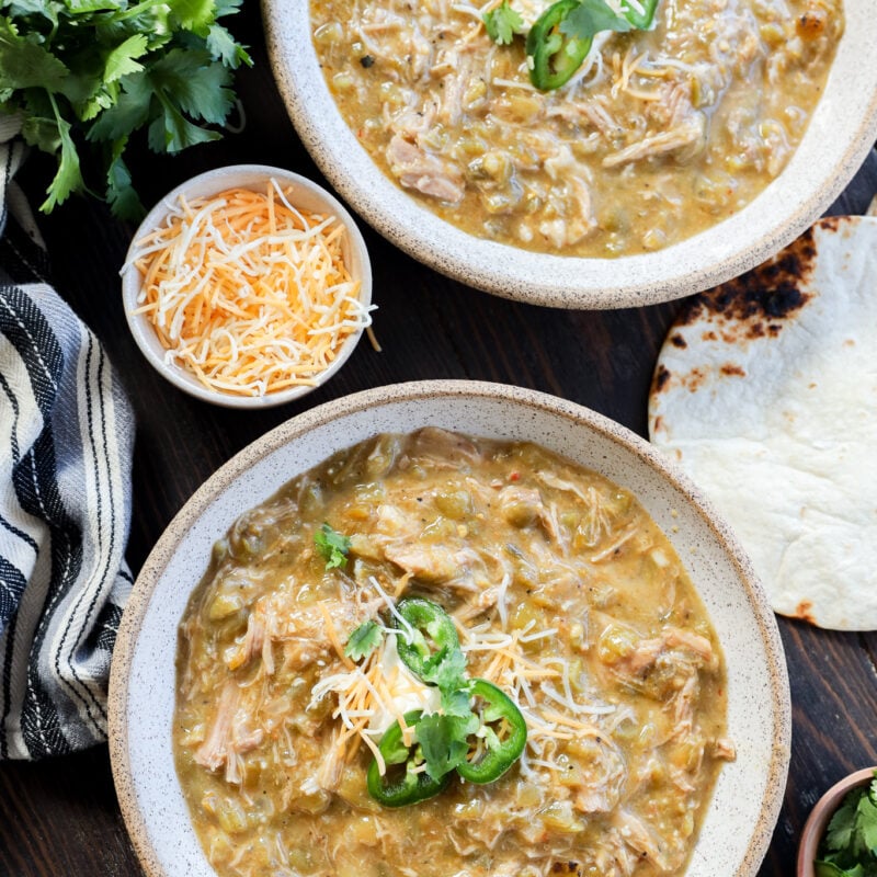 Two bowls of pork green chile stew topped with shredded cheese and sliced jalape&ntilde;os, surrounded by cilantro, extra cheese, a striped towel, and a tortilla on a dark wooden table.