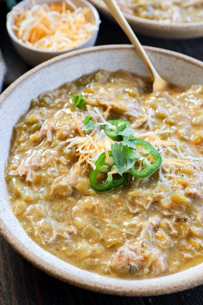 A bowl of pork green chile stew topped with shredded cheese, sliced jalapeños, and cilantro, with a gold spoon; a small bowl of extra shredded cheese sits in the background.