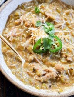 A bowl of pork green chile stew with shredded meat, garnished with sliced jalapeños, shredded cheese, and cilantro, with a spoon resting inside.